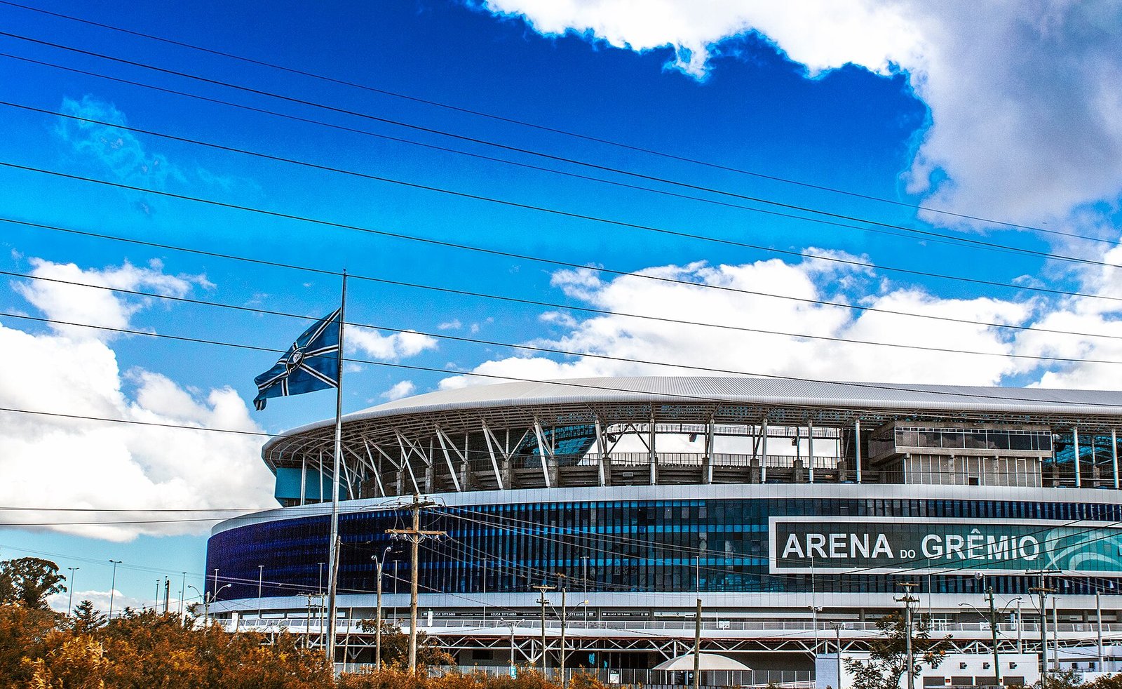 Arena do Grêmio com a bandeira do clube em primeiro plano e o letreiro do estádio na fachada, em Porto Alegre