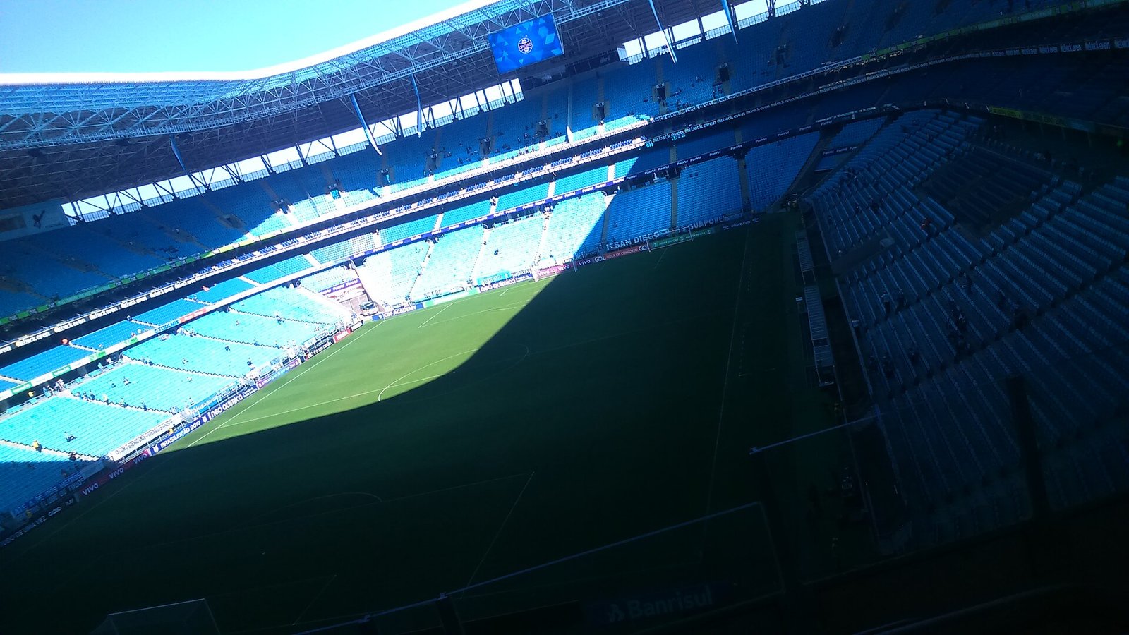 Interior vazio da Arena do Grêmio, estádio do Grêmio em Porto Alegre, com o gramado parcialmente na sombra e as arquibancadas azuis